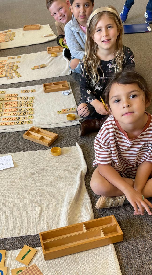 A group of young students sitting on the floor in a classroom, participating in a collaborative Montessori math activity with number tiles and wooden sorting trays.