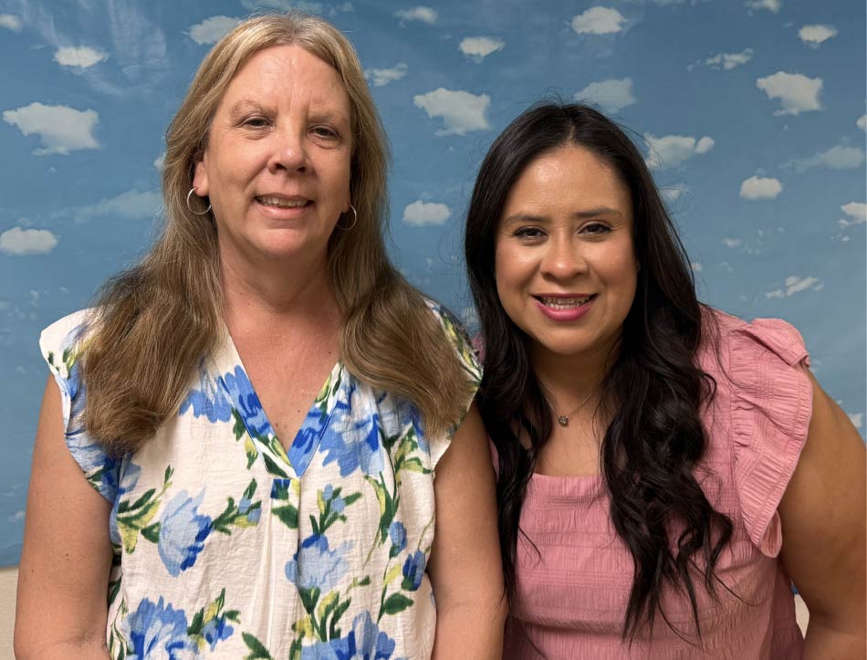 Two smiling female educators from Greenhouse Montessori School posing side-by-side against a blue sky and white cloud backdrop.