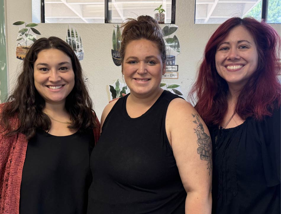 Three smiling female classroom teachers posing together indoors at Greenhouse Montessori School, with plant-themed educational decorations visible on the wall behind them.