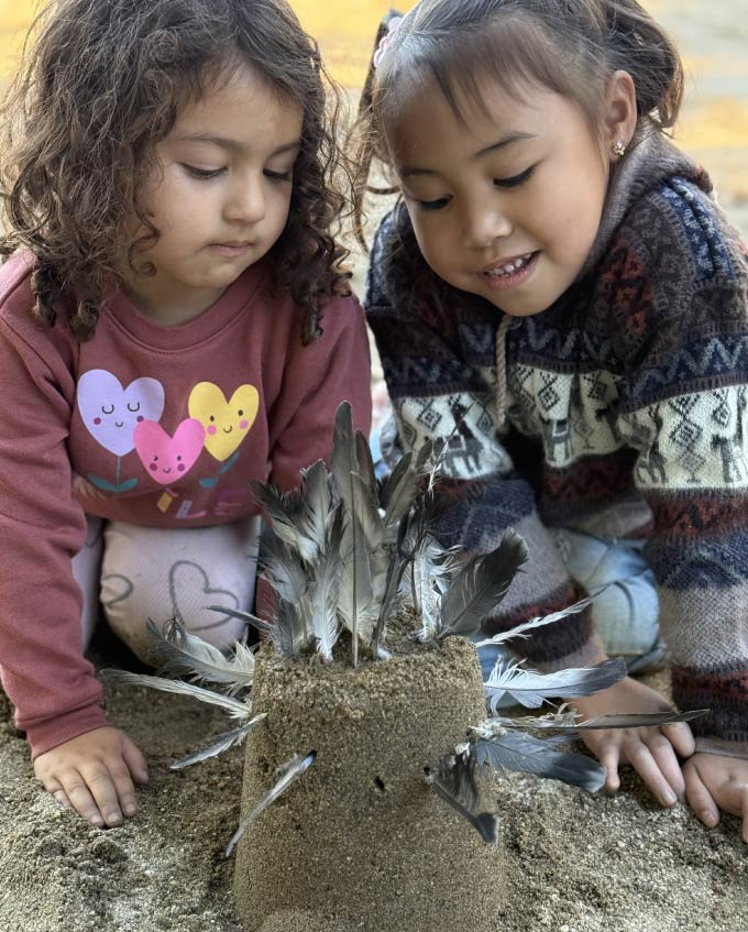 Two young children at Greenhouse Montessori School engaged in a hands-on outdoor sensory activity, focusing on a sand sculpture decorated with feathers.