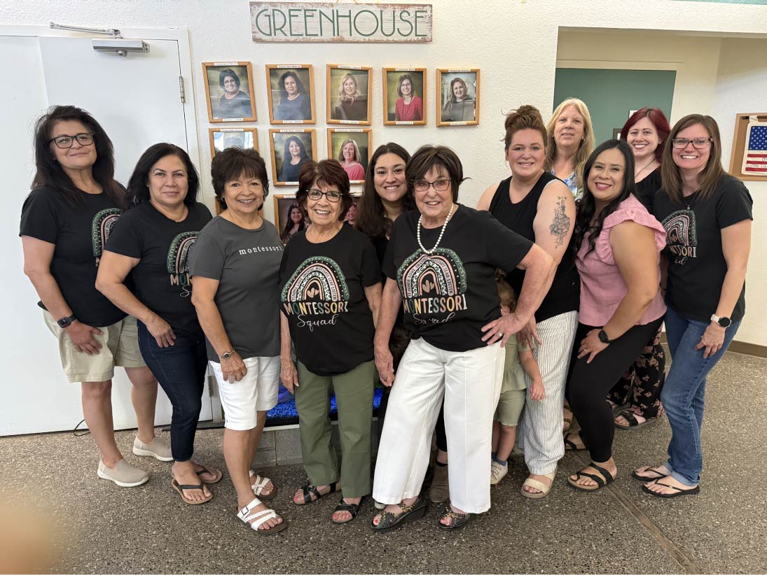 A large group photo of the Greenhouse Montessori School faculty and staff smiling together indoors, with several educators wearing matching black "Montessori Squad" t-shirts beneath a wall display of framed staff portraits.